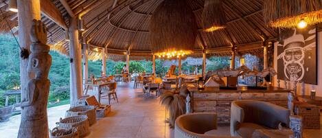 Un área de comedor al aire libre con techo de paja, muebles de madera y pilares de piedra.