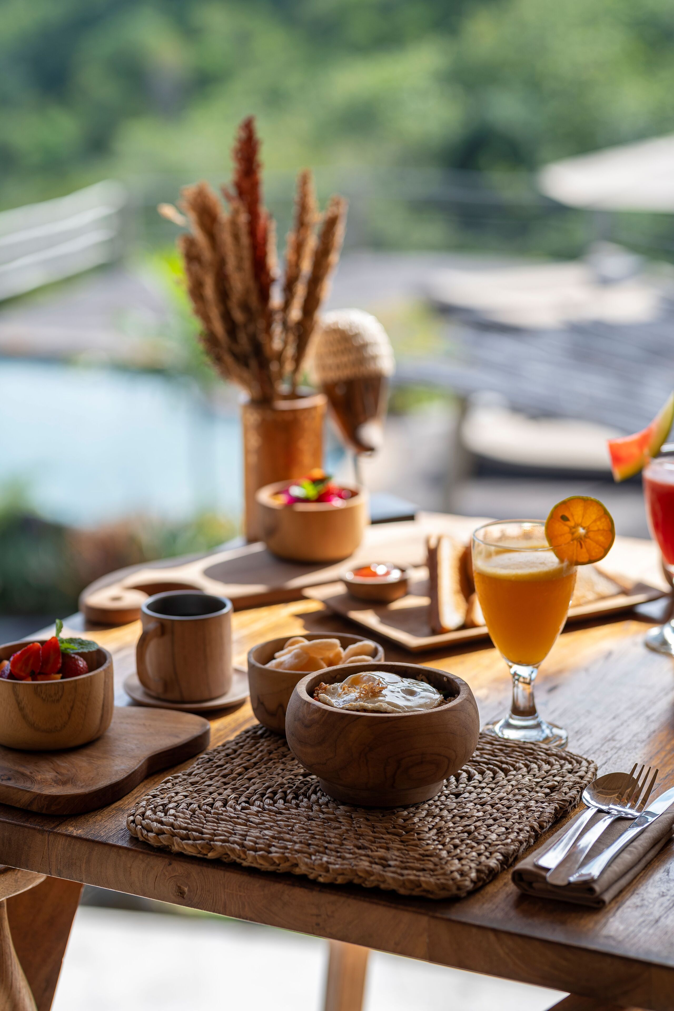 A wooden table with a woven placemat, a glass of orange juice garnished with an orange slice, wooden bowls, and a wooden cup.