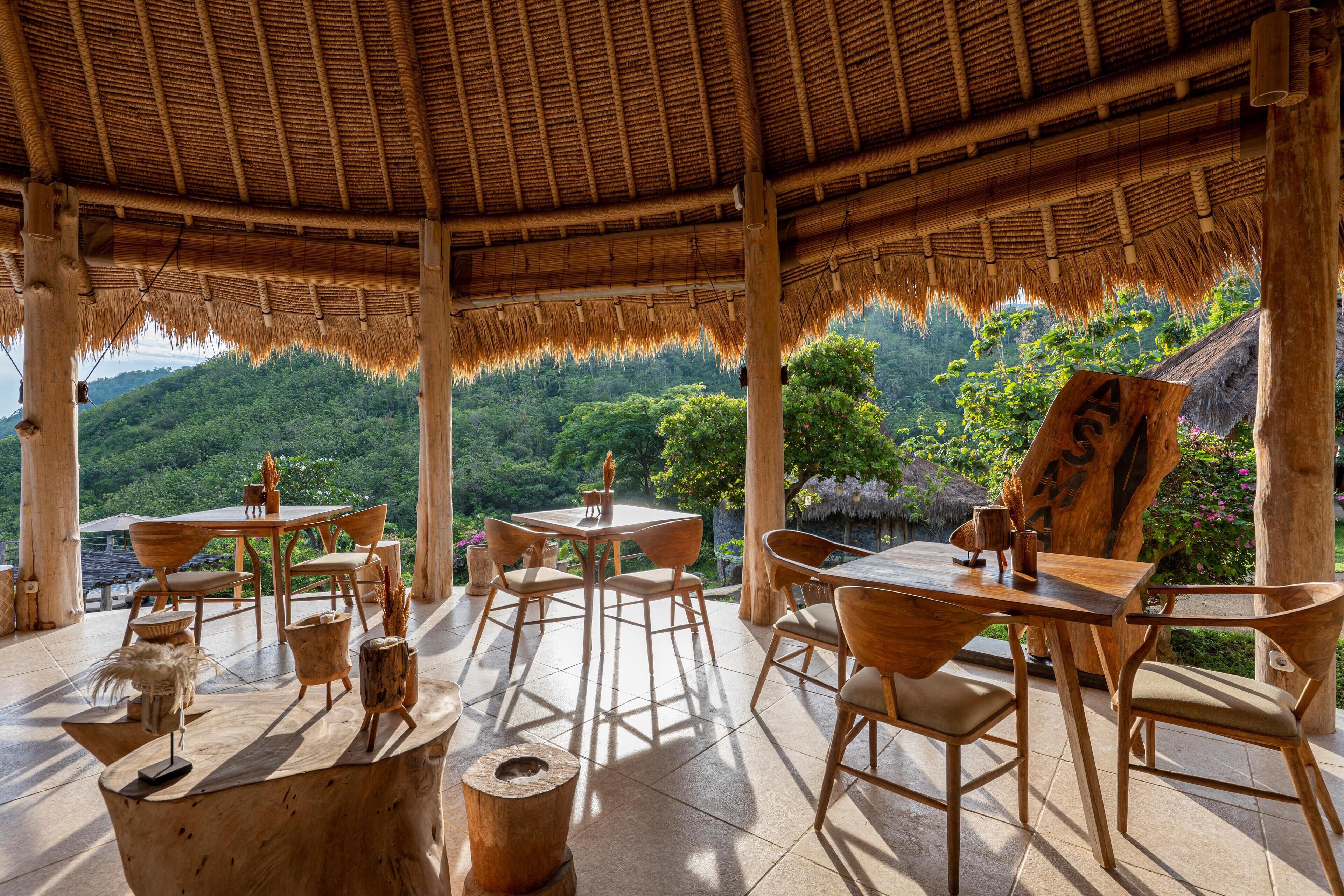 An outdoor dining area with wooden furniture and a thatched roof, overlooking a lush green landscape.