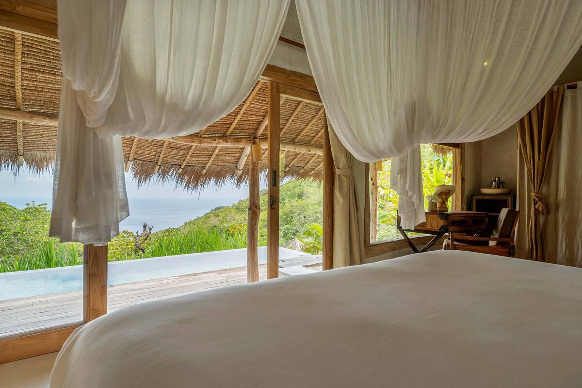 Une chambre dotée d’un grand lit, d’un bureau en bois, d’une chaise et offrant une vue sur un paysage verdoyant et luxuriant ainsi que sur une piscine.