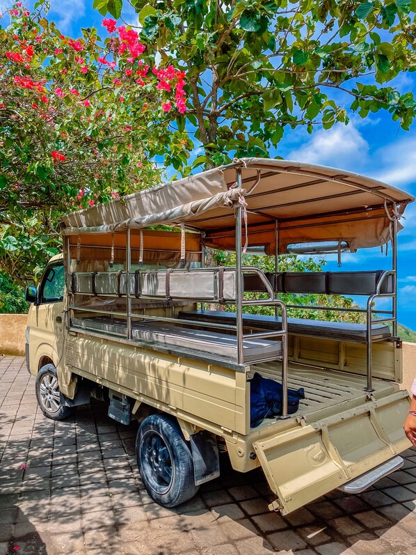 A beige utility vehicle with a canvas roof and metal frame, parked on a paved surface with a tree and sky in the background. - MATHIS Lodge Amed (Karangasem)