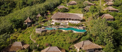 An aerial view of a resort with multiple thatched-roof cottages, a central pool, and lush greenery.
