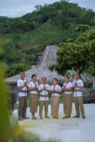 A group of people in traditional attire standing in a courtyard with a thatched-roof structure in the background.