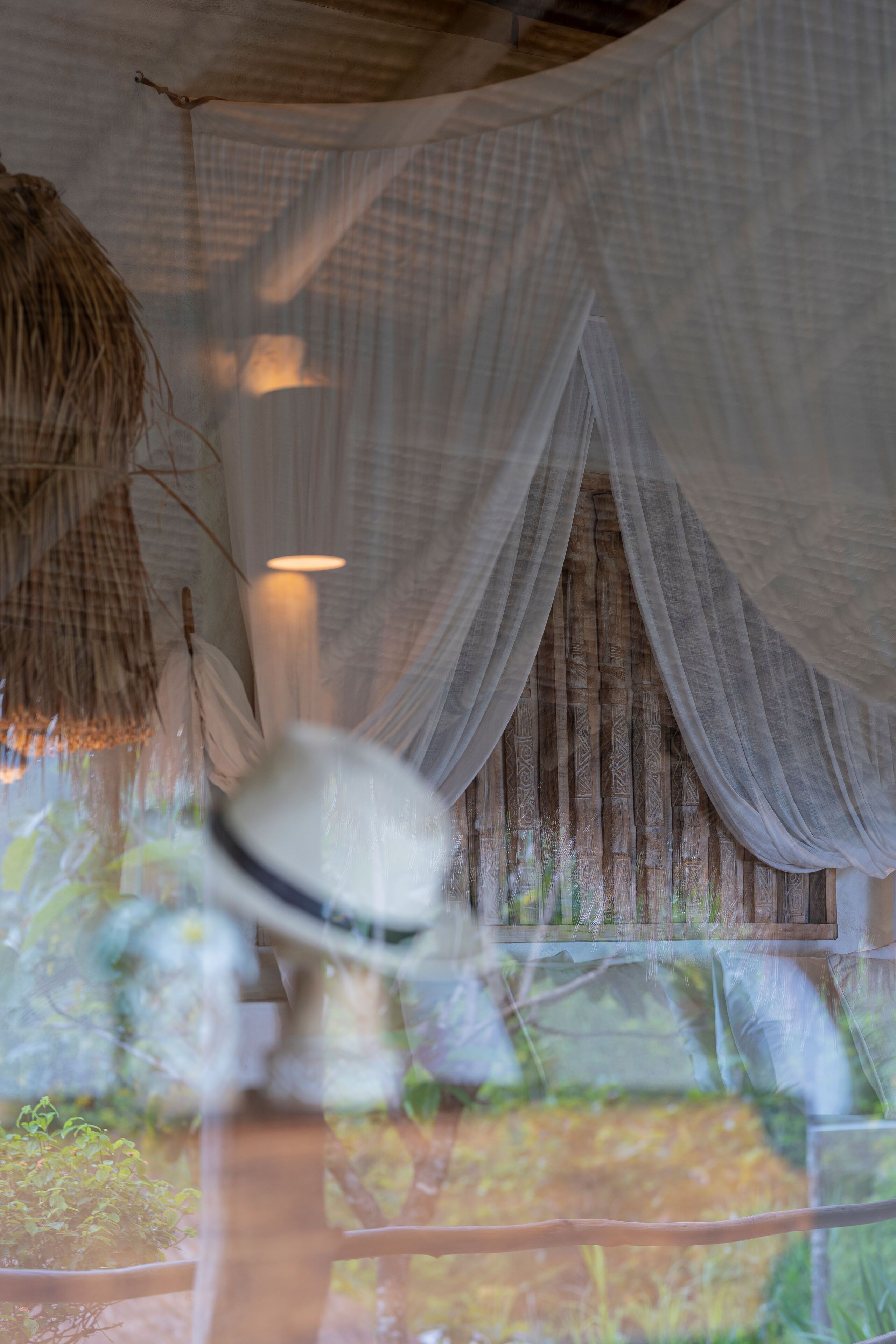 Una habitación con una cama cubierta de sábanas blancas, una lámpara colgante y una ventana con cortinas traslúcidas.