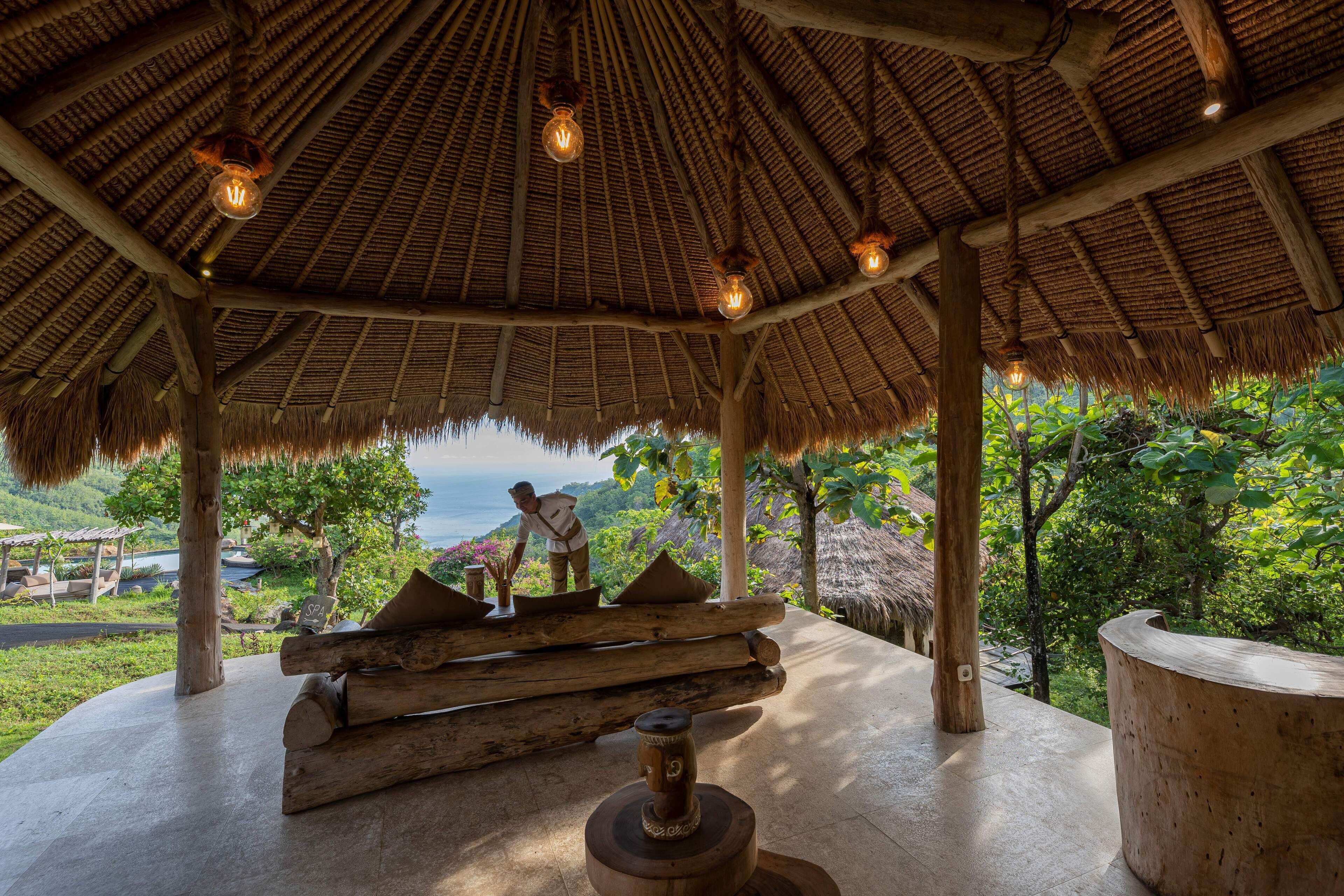 A thatched-roof pavilion with a view of the ocean, wooden logs, and a large wooden sculpture.