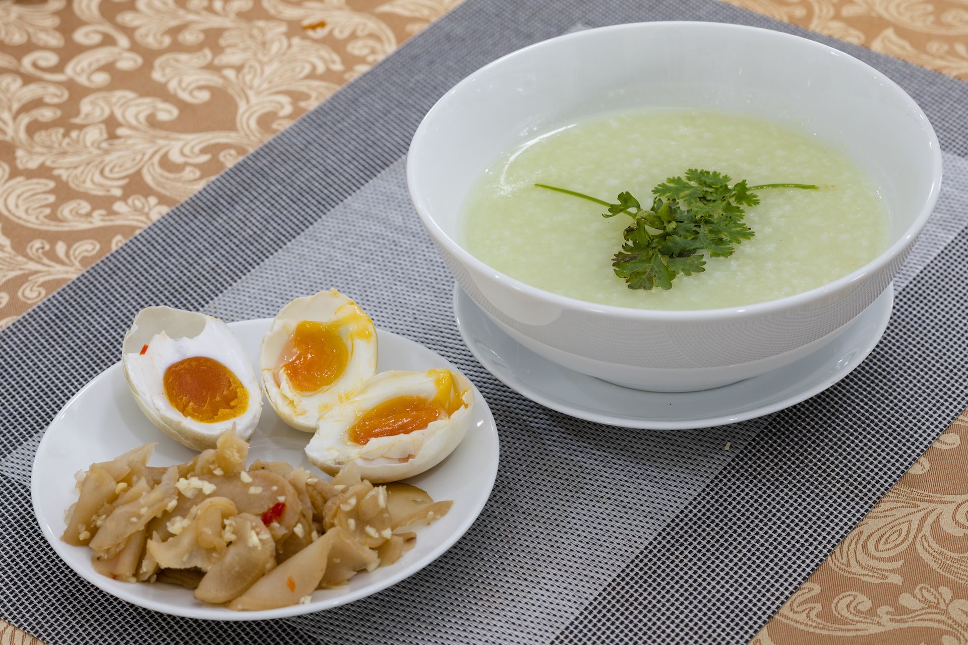 A bowl of green soup garnished with parsley, accompanied by a plate of sliced boiled eggs and a small dish of pickled vegetables.