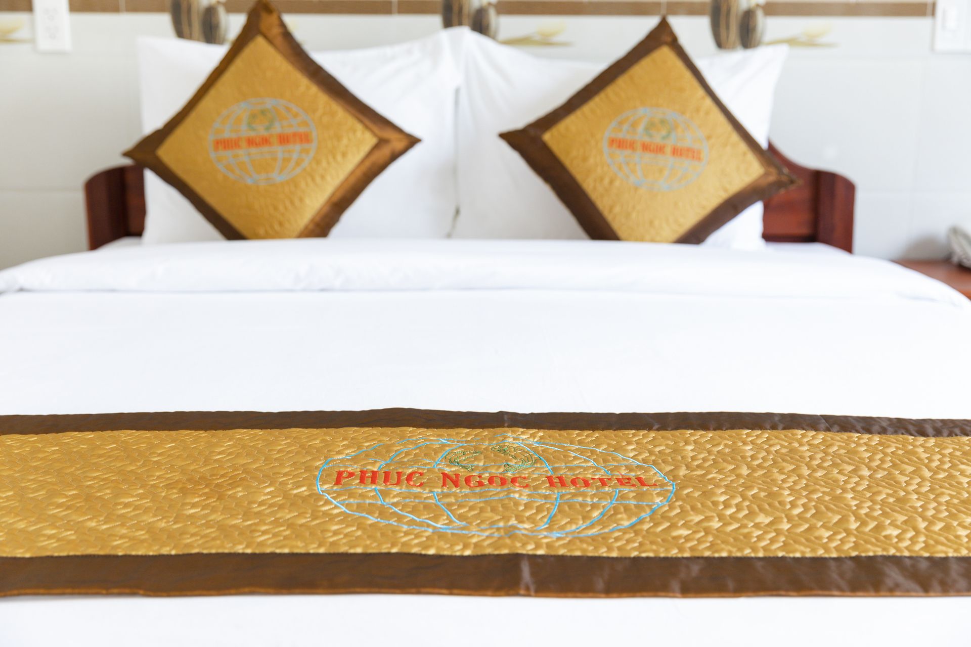 A bed with a gold and brown bedspread, two pillows with a logo, and a white duvet.