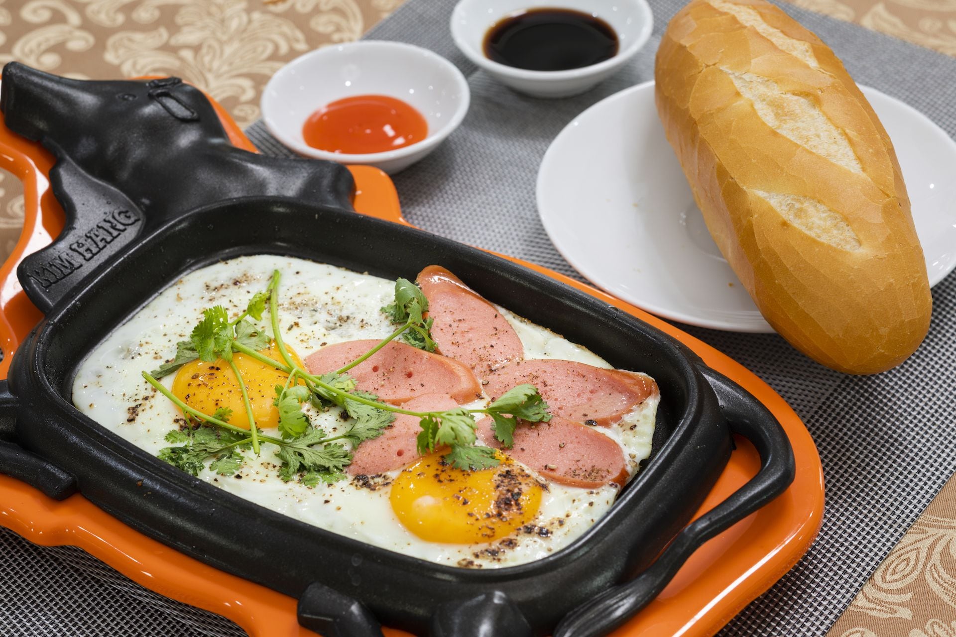 A breakfast dish with eggs, sausage, and herbs, served on a black skillet with orange handles, accompanied by bread and condiments.