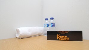Una mesa de madera con una toalla blanca enrollada, dos botellas de agua y una caja negra con el texto "Kamar Eluarga We bring you home, enjoy".