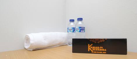 A wooden table with a rolled white towel, two water bottles, and a black box with the text "Kamar Eluarga We bring you home, enjoy."