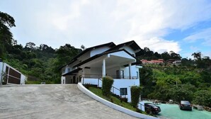 Una casa moderna de dos pisos con un camino curvo, rodeada de exuberante vegetación y colinas.