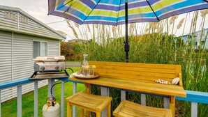 Um deck de madeira com churrasqueira, um banco e uma mesa com bebidas e petiscos.