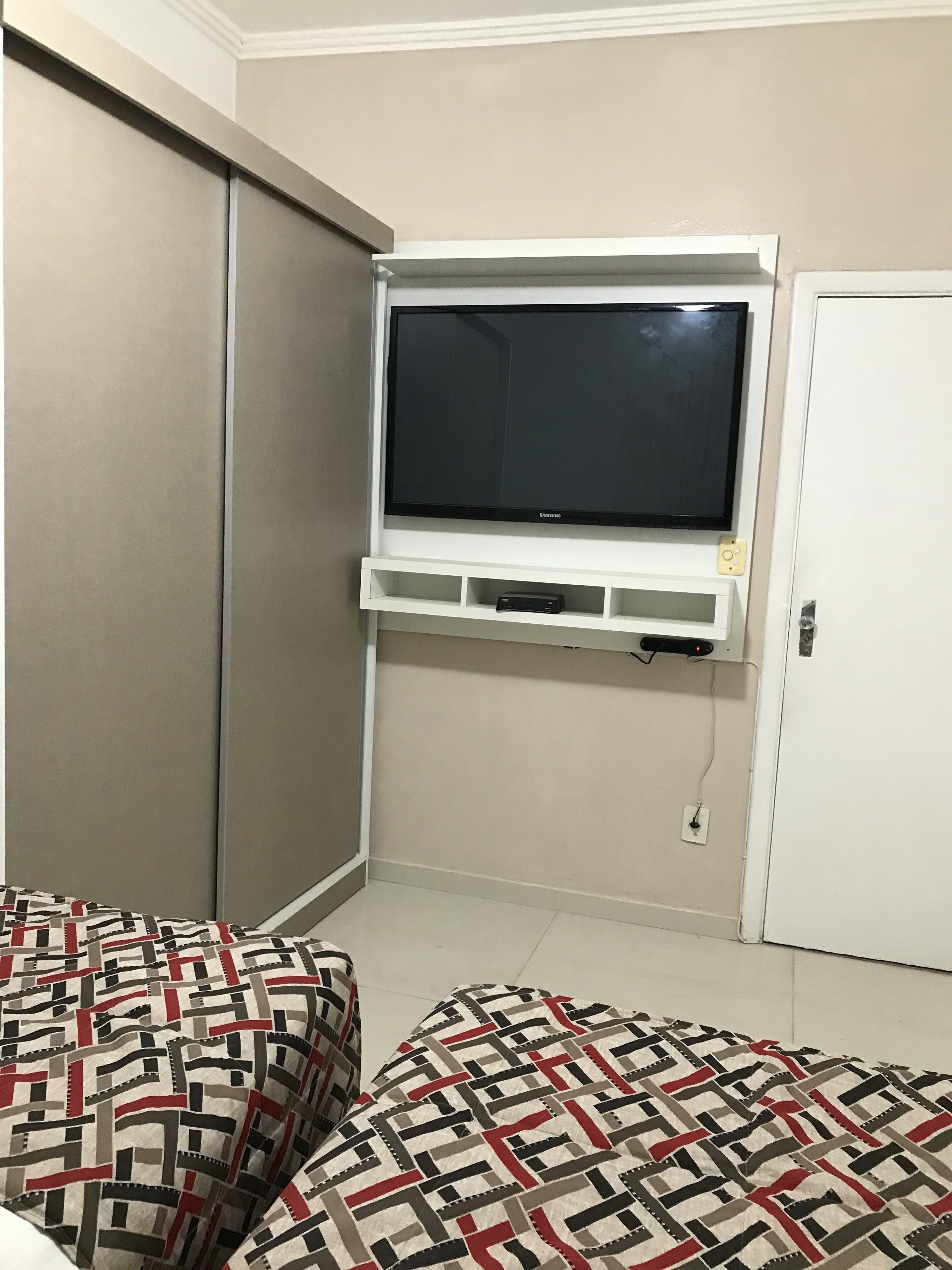 Una habitación con televisión instalada en la pared, una puerta y ropa de cama estampada.