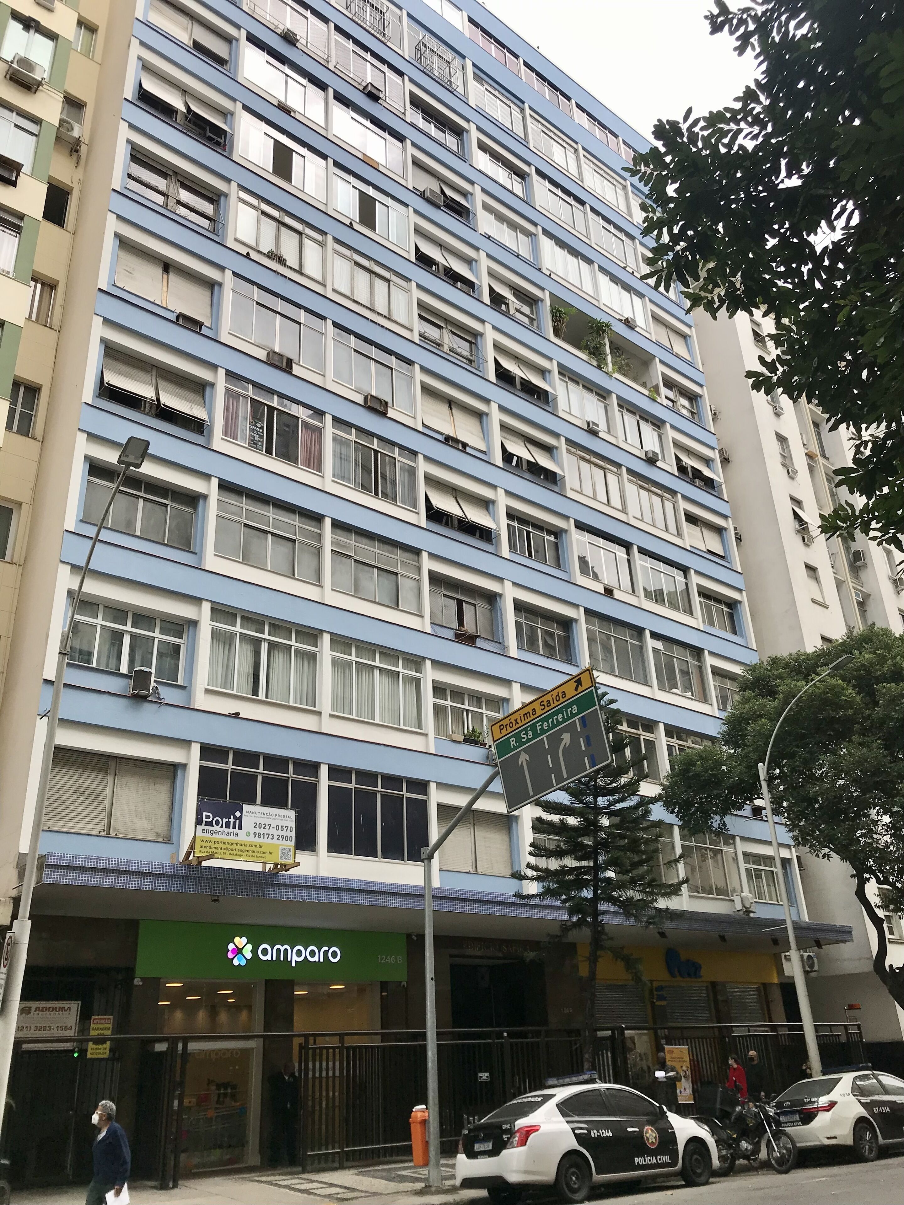 A multi-story building with a blue and white facade, a visible storefront with a green awning, and a sign indicating directions to 'Proxima Saída' and 'R. Sá Ferreira'.