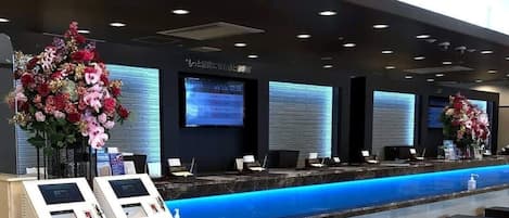 A hotel reception area with a blue-lit reception desk, multiple monitors, and a digital clock displaying 12:58.