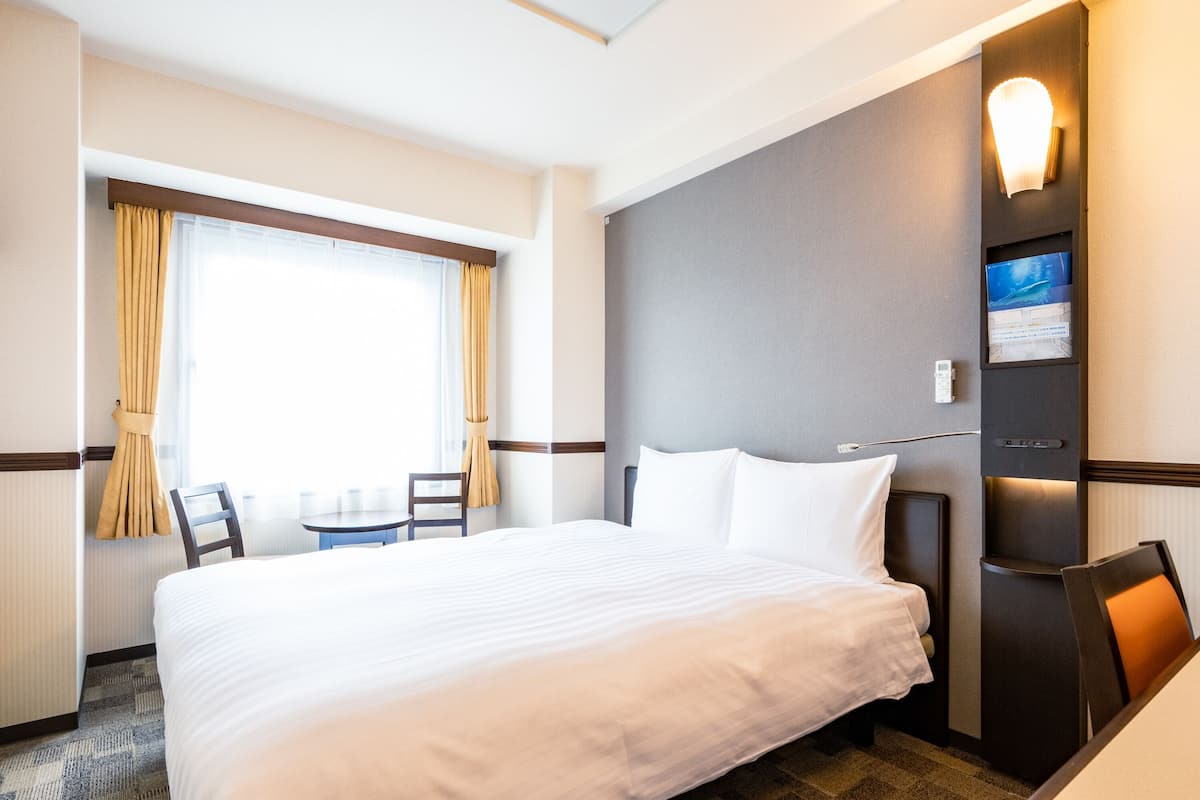 Habitación de hotel con una cama grande, una mesita, dos sillas, una ventana con cortinas y un televisor montado en la pared.