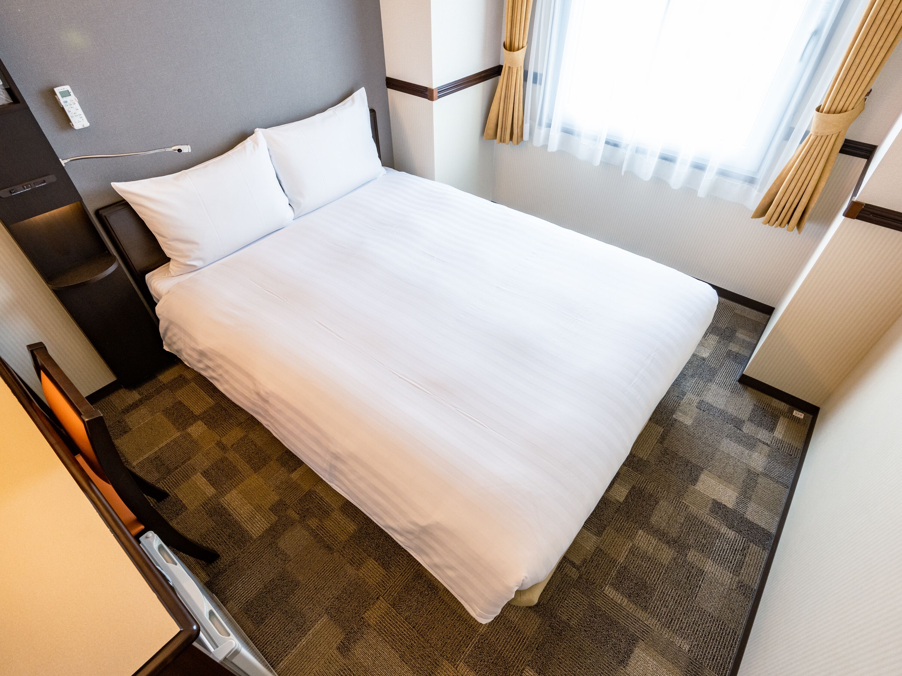 A neatly made bed in a hotel room with a headboard, a bedside table, and a window with curtains.