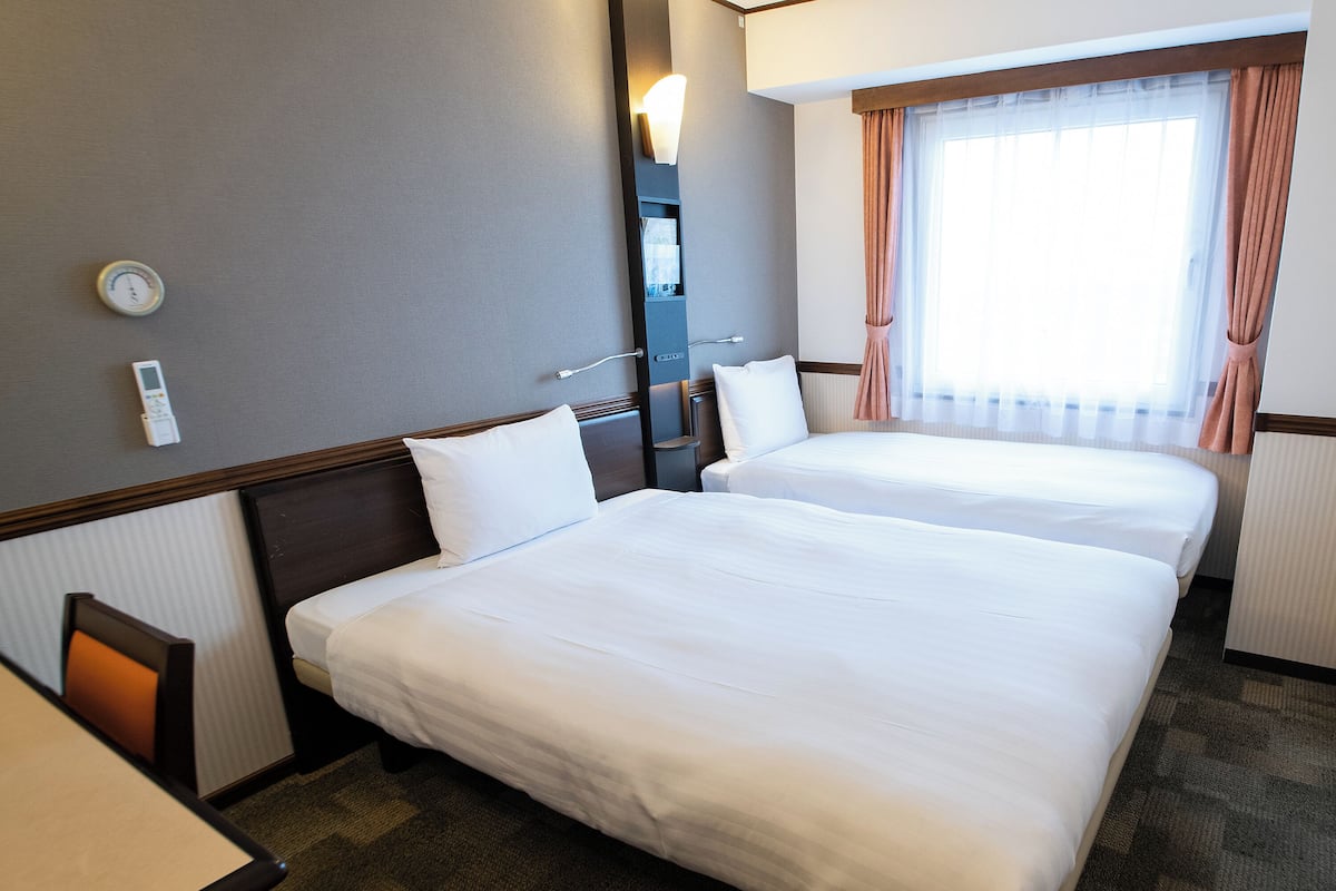 Una habitación de hotel con dos camas, una mesita de noche, una lámpara, una ventana con cortinas y un termostato en la pared.