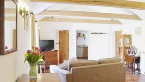 A living room with a sofa, a wooden coffee table, and a TV. There is a kitchen visible through an open door.