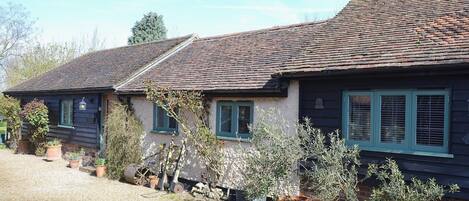 A traditional cottage with a tiled roof, multiple windows with shutters, and potted plants in the garden area.