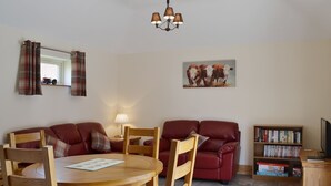 A living room with a wooden dining table, red leather sofas, a wooden bookshelf, and a mounted painting of cows.