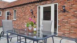 A glass-top table with two chairs, a bottle of wine, and a vase of flowers in front of a brick building with a tiled roof.
