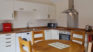 A compact kitchen with white cabinets, a stainless steel range hood, a microwave, a kettle, and a wooden dining table with chairs.