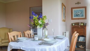 Comedor con una mesa redonda dispuesta para cuatro personas, rodeada de sillas de madera. Un jarrón con flores está sobre la mesa, y se observa una estantería al fondo.