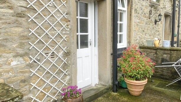Ein Steinhaus mit einer weißen Tür, einem Gitter links und zwei Kübelpflanzen mit Blumen.