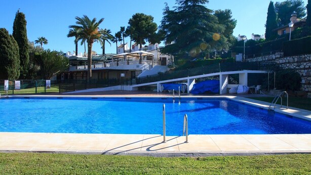 A rectangular swimming pool with clear blue water, surrounded by a well-maintained lawn and various trees. - Cerros del Águila - charming townhouse (Mijas Costa)