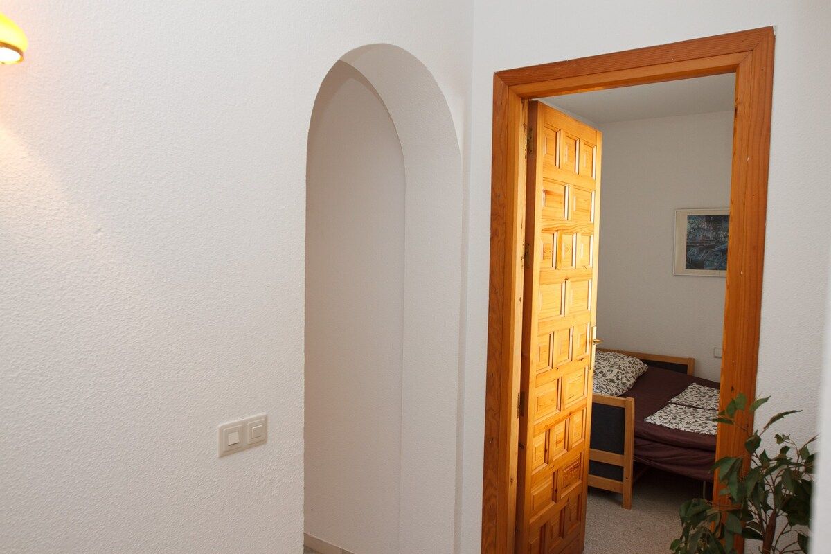 A hallway with a wooden door leading to a bedroom with a bed and a painting on the wall.