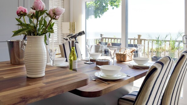 A dining area with a wooden table, white plates, a vase with flowers, and a view of the sea. - 2 bedroom accommodation in Corton, near Lowestoft (Corton, near Lowestoft)