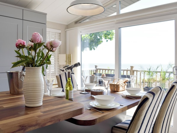 A dining area with a wooden table, white plates, a vase with flowers, and a view of the sea.