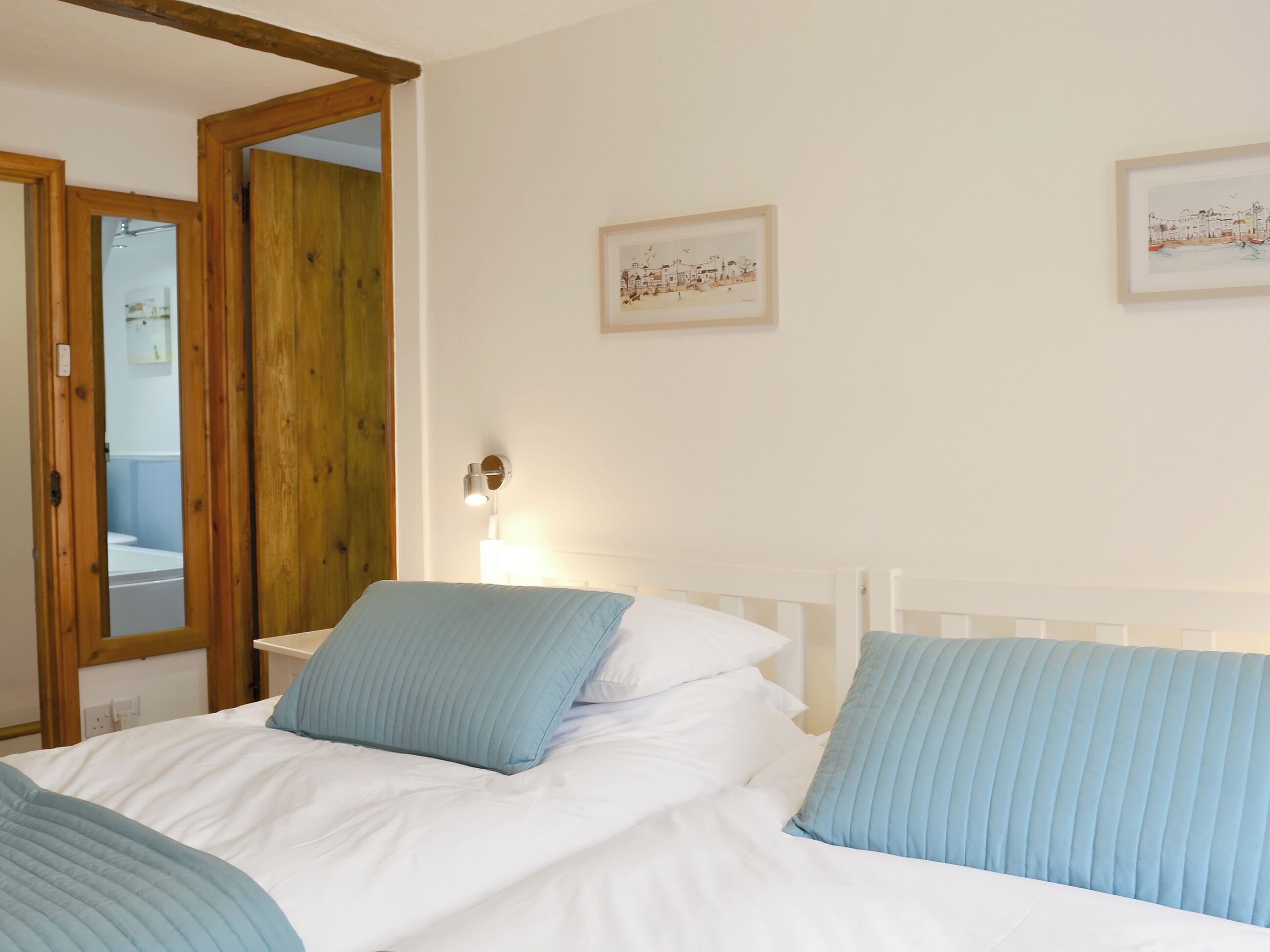 Um quarto com uma cama, uma porta de madeira, uma luminária fixada na parede e duas molduras com imagens.