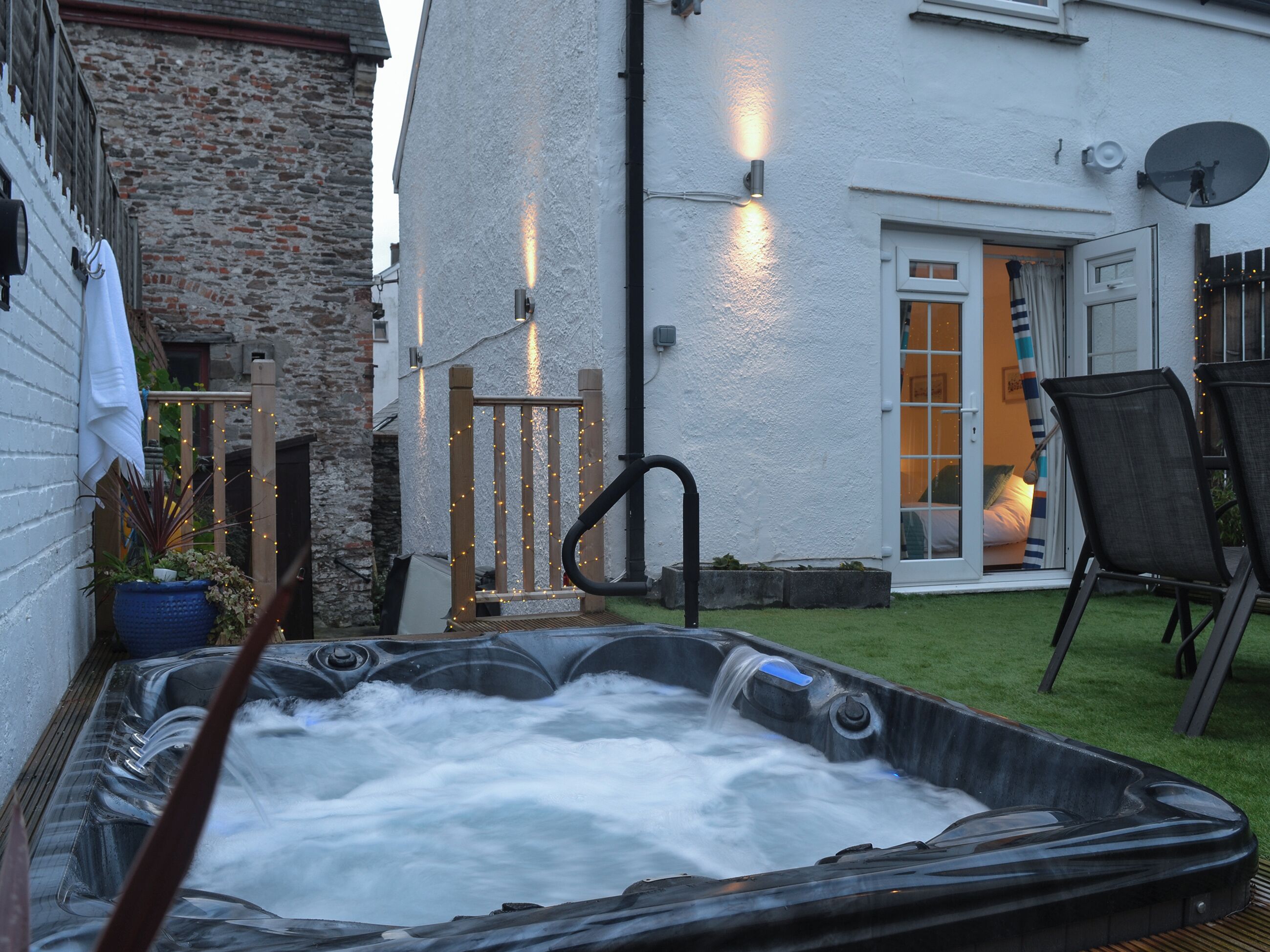 A hot tub in a backyard with a brick wall, wooden deck, and a white building with a door.