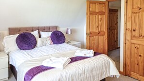 Un lit bien fait, recouvert d’une couverture blanche et violette, avec deux grands oreillers violets et une serviette blanche pliée posée sur le lit.