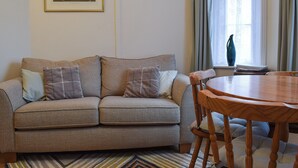 A living room with a sofa, a wooden dining table, and a window with curtains. - 1 bedroom accommodation in Ventnor (Ventnor)