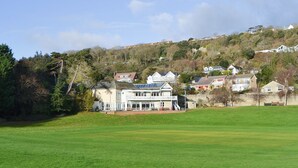 A large, two-story building with a balcony and a solar panel on the roof, surrounded by a well-maintained lawn and a hillside with residential houses. - 1 bedroom accommodation in Ventnor (Ventnor)