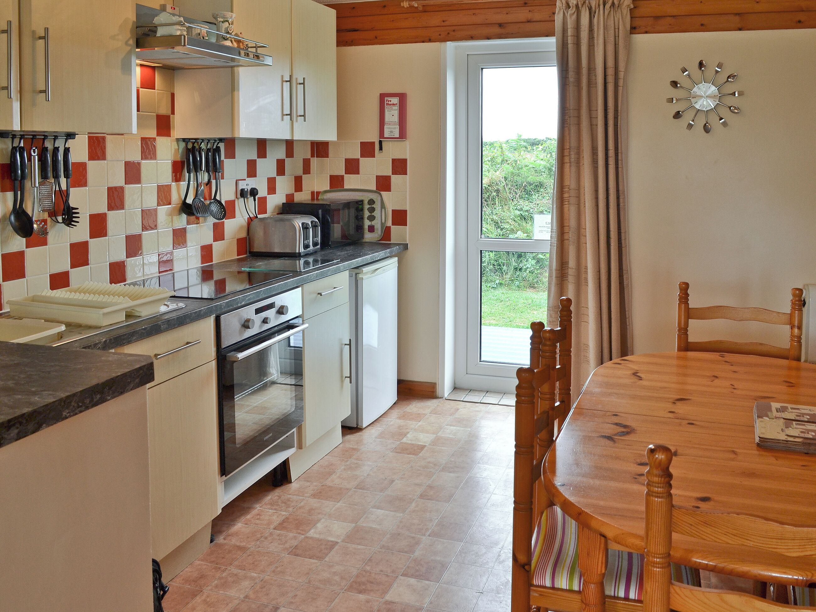 A kitchen with a dining area, featuring a wooden table and chairs, a built-in oven, and a view through a glass door to the outdoors.