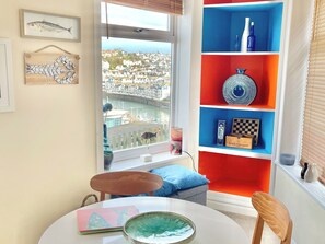 A room with a view of a coastal town, a round table with a bowl, a chair, and a shelf with decorative items.
