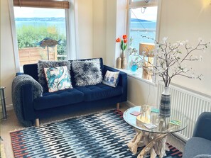 A cozy living room with a dark blue sofa, patterned cushions, a glass coffee table, and a view of the sea through large windows.