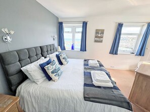 A bedroom with a large bed, a headboard, a window with curtains, a wall-mounted lamp, and a view of the beach.
