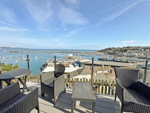 A terrace with wicker furniture overlooking a marina with boats and a coastal town.