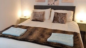Una cama bien hecha con una manta marrón y blanca, dos almohadas y una toalla azul doblada sobre la cama. Hay dos mesitas de noche con lámparas, y tres cuadros enmarcados con una escena de playa en la pared.