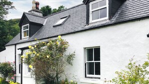 A white house with a black roof, two windows, and a garden with yellow flowers. - 3 bedroom accommodation in Lothmore, Helmsdale (Lothmore, Helmsdale)