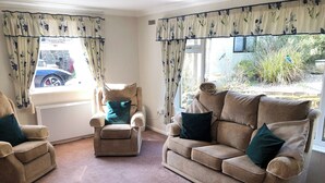 Un salón con sofá color beige, un sillón a juego y un sillón reclinable. Hay cortinas con motivos florales, un radiador y vistas al exterior.