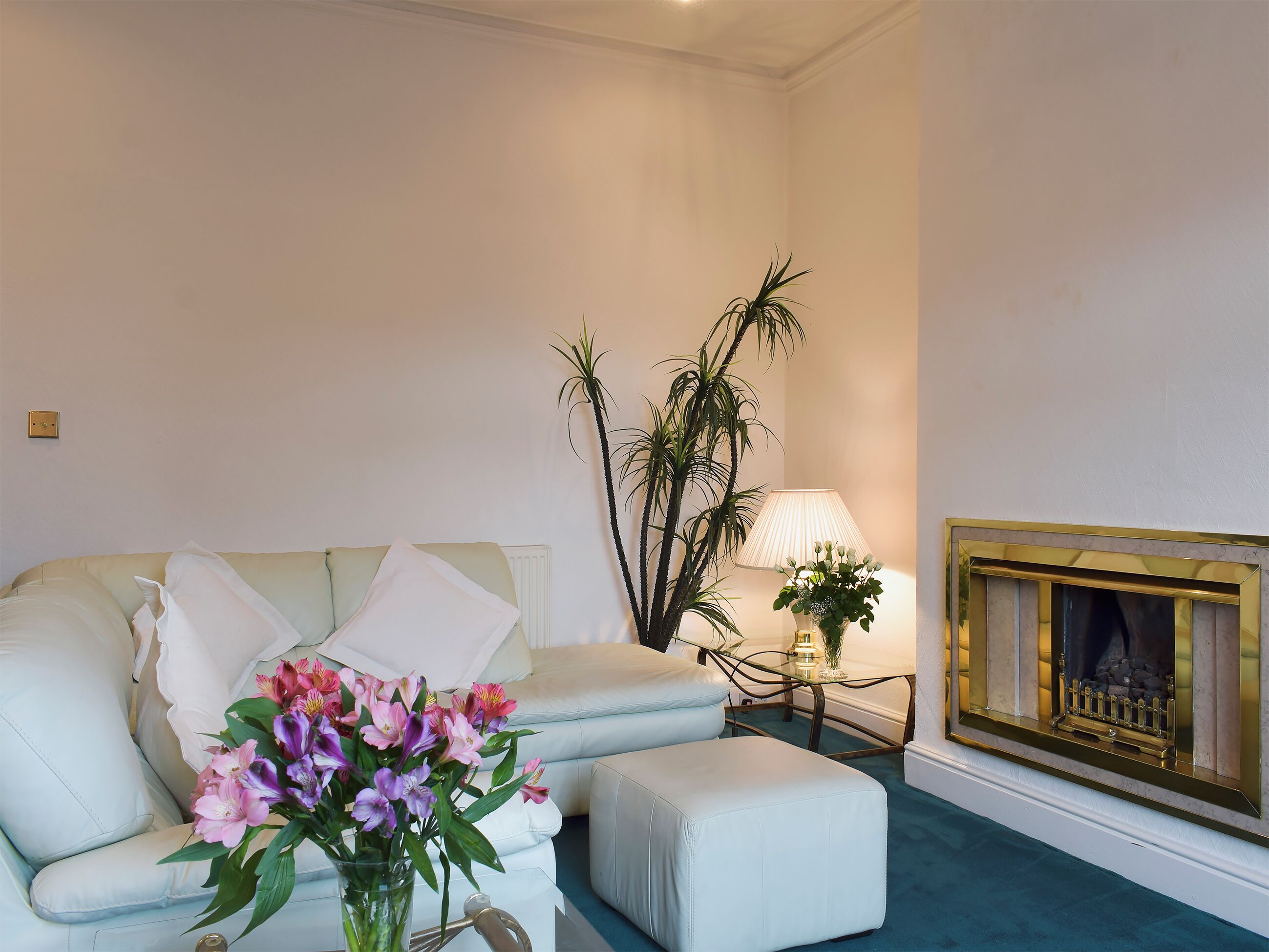 A living room with a white sofa, a small table with a lamp, a fireplace, and a vase of flowers.