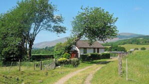 Une maison de campagne, accessible par un chemin de terre, entourée de verdure et avec des collines en arrière-plan.