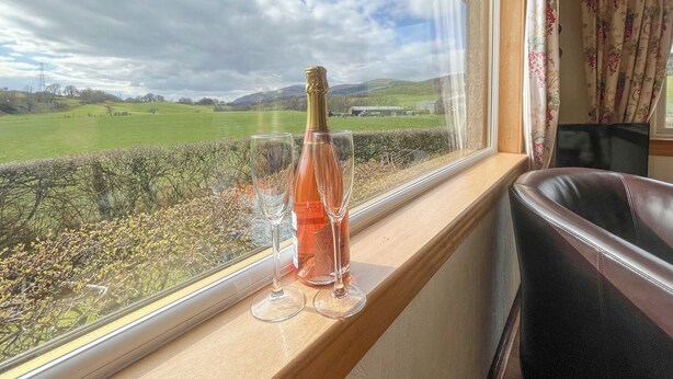 Une fenêtre avec une bouteille de vin rosé et deux verres posés sur un rebord en bois, donnant sur un paysage verdoyant avec des collines et un ciel nuageux.