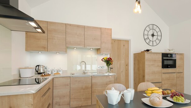 A modern kitchen with wooden cabinets, a built-in oven, a coffee station, and a dining area with a table set for two. - The Old Meeting Hall (King&rsquo;s Somborne, near Winchester)